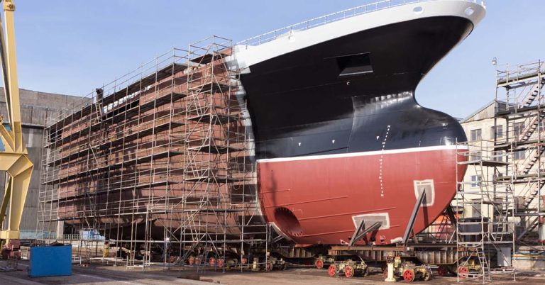Large ship hull under construction with red and black paint, metal scaffolding, and welding in progress at shipyard